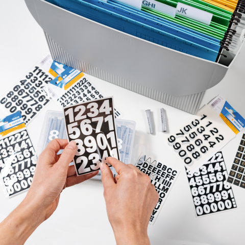 Hände halten eine schwarze Tafel mit weißen Zahlen. Im Hintergrund sind zusätzliche Zahlen und Buchstaben sowie ein Ordnungssystem zu sehen.