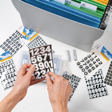 Hände halten eine schwarze Tafel mit weißen Zahlen. Im Hintergrund sind zusätzliche Zahlen und Buchstaben sowie ein Ordnungssystem zu sehen.