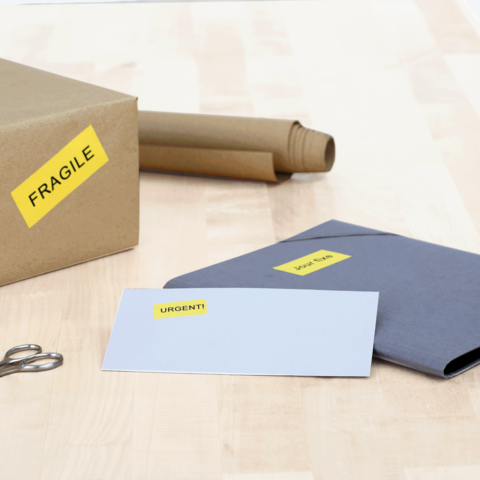 Ein Karton mit dem Aufkleber "FRAGILE" steht neben einem grauen Umschlag mit der Aufschrift "DRINGEND!" und einer Schere.