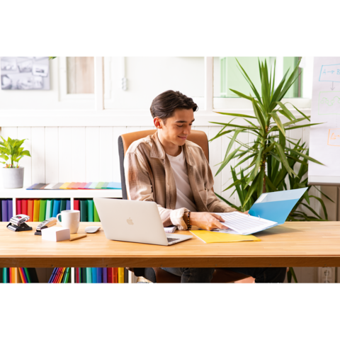 Ein junger Mann sitzt an einem Schreibtisch, arbeitet mit einem Laptop und blättert durch Dokumente in einer Ordnermappe. Pflanzen und farbige Akten sind im Hintergrund.