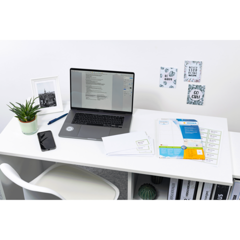 Ein Schreibtisch mit einem Laptop, einem Smartphone, Pflanzen und verschiedenen Dokumenten. An der Wand hängen dekorative Notizen.