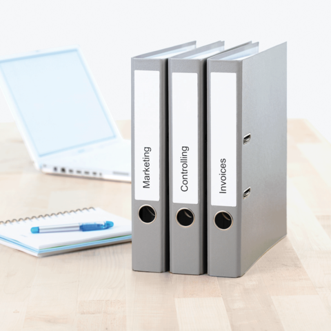 Drei graue Ordner mit Beschriftungen "Marketing", "Controlling" und "Rechnungen" stehen neben einem Laptop und einem Notizblock.