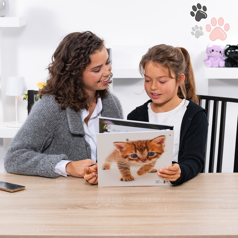 Eine Frau und ein Mädchen sitzen an einem Tisch und schauen sich gemeinsam ein Bilderbuch über Katzen an.