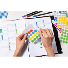 Hände halten ein Blatt mit bunten Aufklebern über einem offenen Planer, um Termine oder Aufgaben zu kennzeichnen.