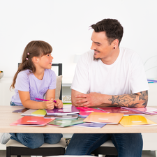 Ein Mann und ein Mädchen sitzen an einem Tisch, umgeben von bunten Farbmusterkarten, während sie miteinander sprechen.