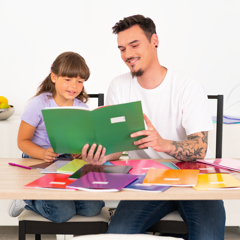 Ein Mann und ein Mädchen sitzen an einem Tisch, umgeben von bunten Heften und Schreibmaterialien, während sie gemeinsam ein Heft betrachten.