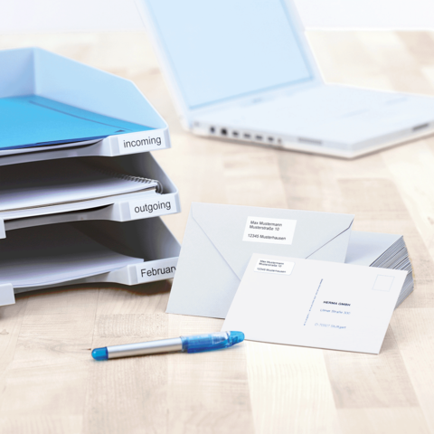 Ordner mit beschrifteten Fächern für eingehende und ausgehende Dokumente, daneben Umschläge und ein blauer Stift auf einem Schreibtisch.