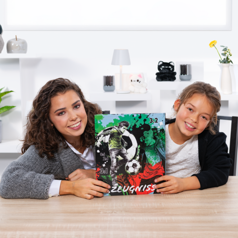 Zwei Mädchen halten ein Fußballzeugnis mit einer Grafik und der Punktzahl 3:2. Sie sitzen an einem Tisch in einem hellen Raum.
