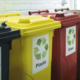 Drei Mülltonnen in verschiedenen Farben mit Recycling-Symbolen: Gelb für Plastik, Rot für Glas und Schwarz für Restmüll.