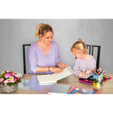 Eine Frau und ein Mädchen sitzen an einem Tisch und betrachten gemeinsam ein Buch. Auf dem Tisch liegen mehrere Schreibutensilien und ein Blumenstrauß.