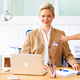 Eine blonde Frau in einem beigen Blazer sitzt an einem Schreibtisch mit einem Laptop und lächelt. Sie hat ein Namensschild um den Hals.