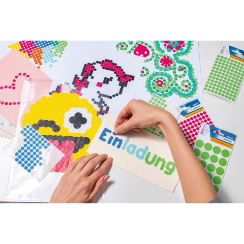 Bunte Bastelmaterialien liegen auf einem Tisch. Eine Hand platziert Punkte auf eine Einladungskarte mit dem Wort "Einladung".
