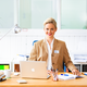 Eine Frau mit blonden Haaren sitzt an einem Schreibtisch, lächelt und trägt einen blauen Blazer. Vor ihr steht ein Laptop sowie Schreibutensilien.