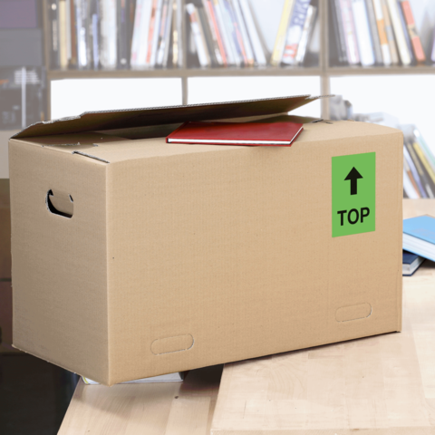 Kartonbox auf einem Tisch, mit einem roten Heft und einem grünen "TOP"-Schild auf der Seite. Hintergrund mit Bücherregalen.