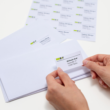 Ein junger Mann sitzt an einem Tisch, hält ein Blatt mit Aufklebern in der Hand und lächelt. Auf dem Tisch sind ein Laptop und eine Verpackung zu sehen.