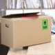 Kartonbox auf einem Tisch, mit einem roten Heft und einem grünen "TOP"-Schild auf der Seite. Bücherregal im Hintergrund.