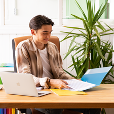 Ein junger Mann sitzt an einem Schreibtisch, arbeitet mit einem Laptop und blättert durch Dokumente in einer Ordnermappe. Pflanzen und farbige Aktenordner sind im Hintergrund.