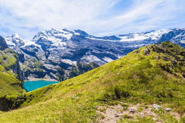 Panoramablick auf sanfte grüne Hügel und schneebedeckte Berge, umgeben von klarem Wasser und strahlend blauem Himmel.