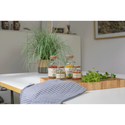 Glasbehälter mit Gewürzen und frischen Kräutern auf einem Holzbrett, daneben ein kariertes Geschirrtuch und eine Topfpflanze im Hintergrund.