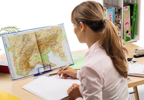 Frau, die vor einem geöffneten Atlas an einem Schreibtisch sitzt.