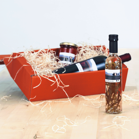 Ein orangefarbener Geschenkkorb mit einer Flasche Likör, einem Glas Marmelade und einer Flasche Wein, umgeben von Holzwolle.