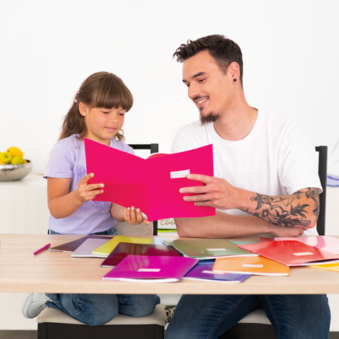 Ein Mann und ein Mädchen sitzen an einem Tisch und lesen gemeinsam in einem bunten Heft. Mehrere Notizhefte liegen verstreut auf dem Tisch.