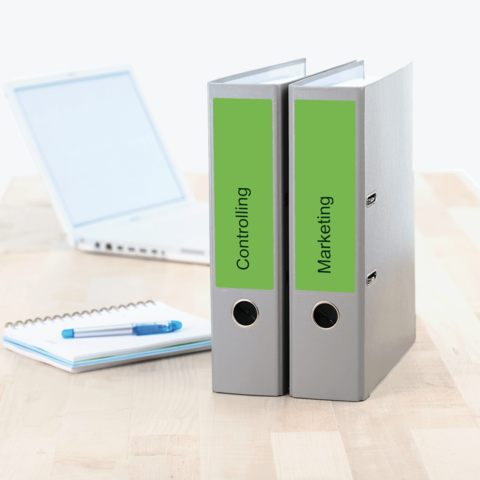Zwei Aktenordner mit den Beschriftungen "Controlling" und "Marketing" stehen auf einem Tisch neben einem Laptop und einem Notizblock.
