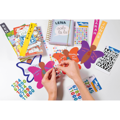 Hände, die bunte Aufkleber auf Schmetterlingsformen anbringen, umgeben von Notizbüchern, Aufklebern und anderen Bastelmaterialien.