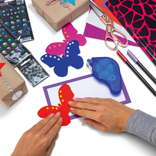 Hände formen bunte Schmetterlinge aus Papier, umgeben von Bastelmaterialien wie Geschenken, Aufklebern und Stiften.
