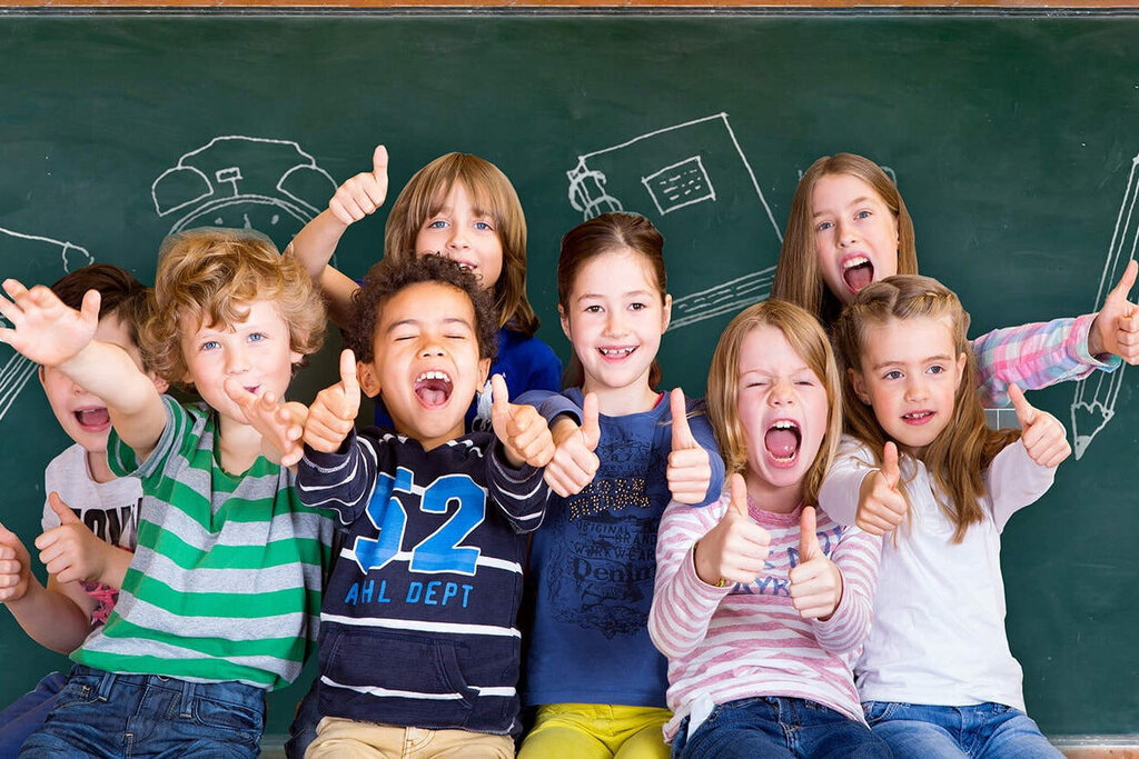 Eine Gruppe von Kindern steht vor einer Tafel, lächelt und posiert fröhlich. Im Hintergrund sind Zeichnungen von Schulmaterialien zu sehen.