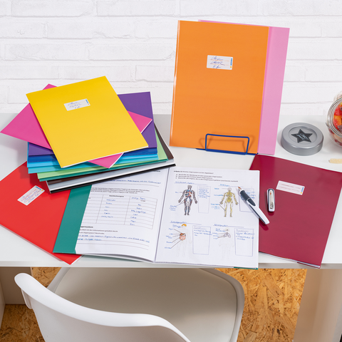 Bunte Ordner und Dokumente auf einem Schreibtisch, daneben ein Glas mit Süßigkeiten und Bürobedarf wie ein Hefter und ein Stift.