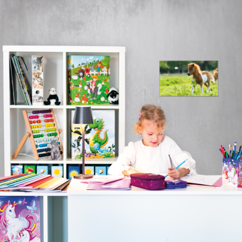 Ein Mädchen sitzt an einem Schreibtisch, malt und schreibt in ein Heft. Im Hintergrund sind Regale mit Spielzeug und Kunstmaterialien.