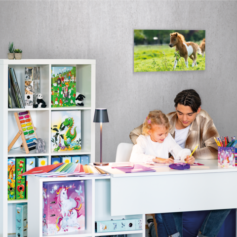 Ein Mädchen sitzt an einem Schreibtisch, malt und schreibt in ein Heft. Im Hintergrund sind Regale mit Spielzeug und Kunstmaterialien.