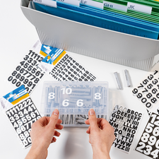 Hände halten eine transparente Box mit Zahlen und Buchstabenaufklebern, umgeben von weiteren Aufklebern und einem Ordner mit farbigen Trennblättern.
