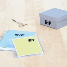 Ein blauer Notizblock, eine gelbe CD-Hülle mit dem Logo "E!F", eine Schere und eine graue Geschenkbox auf einem Holztisch.