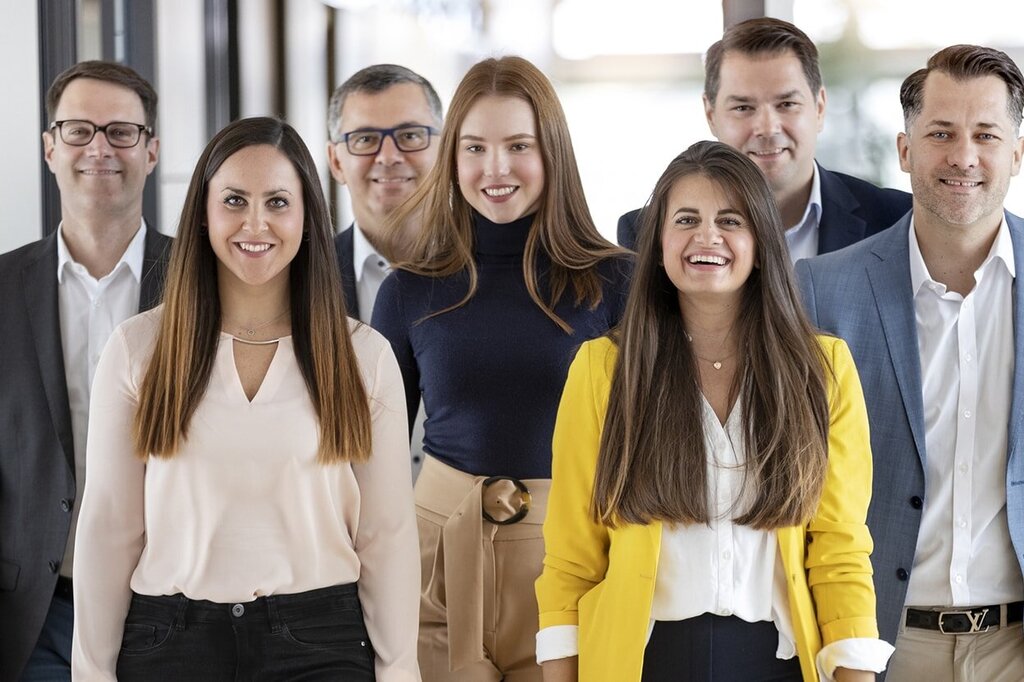 Eine Gruppe von sieben Personen steht in einem Flur, lächelt und trägt formelle Kleidung; einige haben Blusen und Anzüge an.