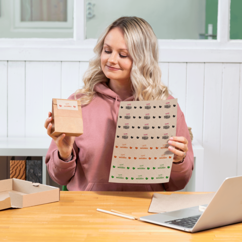 Eine Frau mit blonden, lockigen Haaren hält eine kleine Box und ein Blatt mit Aufklebern in der Hand, während sie an einem Tisch sitzt.