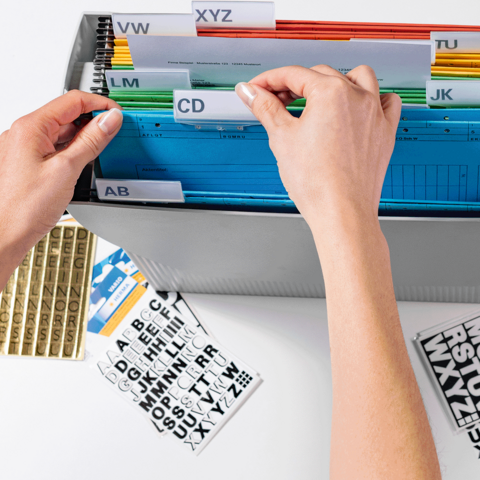 Eine Hand sortiert bunte Registerkarten in einem Ordner, während daneben Etiketten mit Buchstaben liegen.