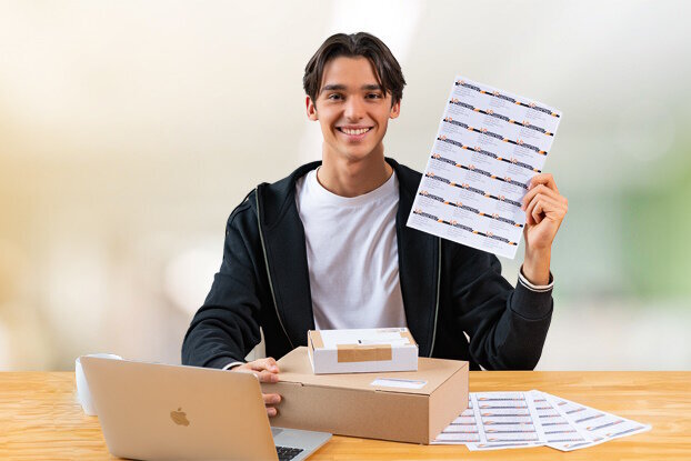 Ein junger Mann sitzt an einem Tisch mit einem Laptop und hält ein Blatt mit Aufklebern in der Hand.