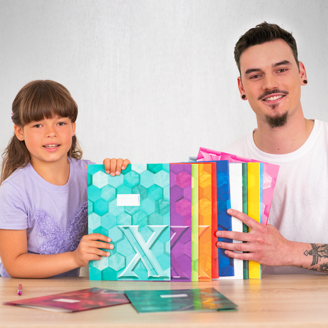 Ein Kind und ein Erwachsener halten bunte, geometrisch gestaltete Ordner und zeigen auf einen der Ordner. Sie sitzen an einem Tisch.