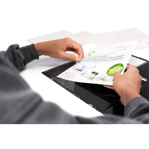 Eine Hand hält einen Gutschein, während sie in einem schwarzen Ordner blättert. Im Hintergrund sind weitere Dokumente sichtbar.