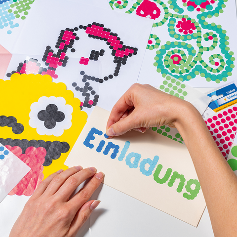 Eine Hand platziert bunte Punkte auf ein Einladungskarten-Design mit einem Monster und verschiedenen bunten Mustern auf dem Tisch.