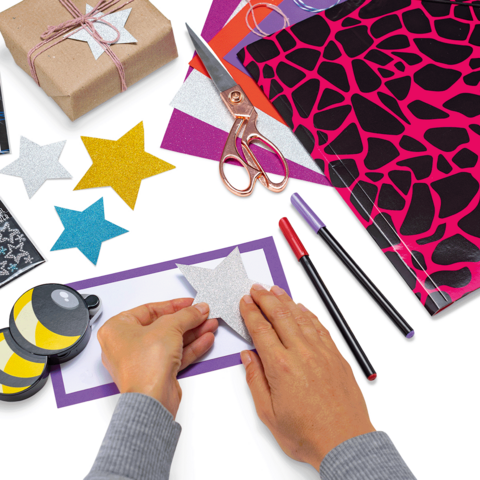 Hände, die einen funkelnden Stern auf ein buntes Blatt Papier kleben, umgeben von Bastelmaterialien wie Scheren, Stiften und Geschenkpapier.