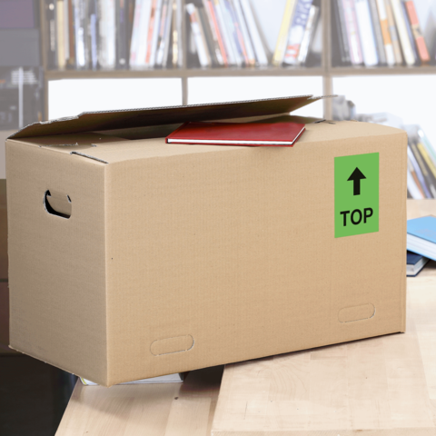 Kartonbox auf einem Tisch, mit einem roten Heft oben und einem grünen "TOP"-Schild. Im Hintergrund sind Bücherregale sichtbar.