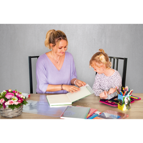 Eine Frau und ein Mädchen sitzen an einem Tisch, schauen in ein Buch und interagieren. Auf dem Tisch liegen verschiedene Schreibmaterialien und Blumen.