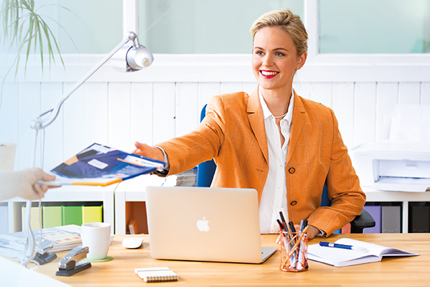 Zwei Personen sitzen an einem Tisch und präsentieren Etiketten. Links ein Mann mit Etikettenblättern, rechts eine Frau mit einem Laptop und Etiketten.