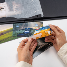 Eine Person hält eine Packung Fotohüllen und betrachtet mehrere Fotos auf einem Tisch, während ein Fotoalbum im Hintergrund sichtbar ist.