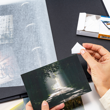 Eine Person hält eine Fotografie in der Hand, während sie ein Fotoalbum mit weiteren Bildern und einer Packung Fotohüllen betrachtet.