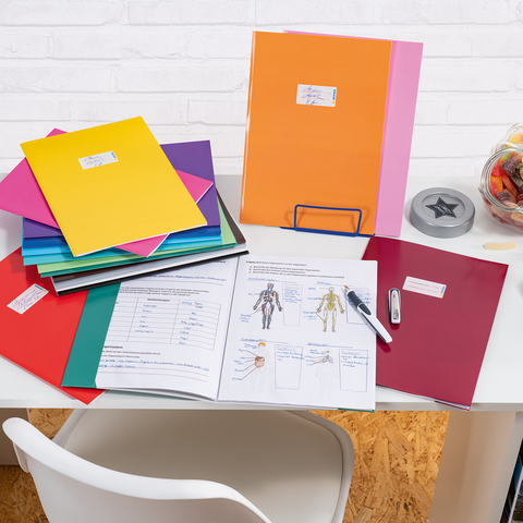 Bunte Ordner und Unterlagen auf einem Schreibtisch, daneben ein Glas mit Süßigkeiten und Bürobedarf wie einen Hefter und einen Stift.
