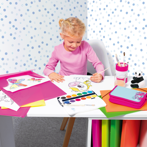 Ein kleines Mädchen sitzt an einem Tisch und malt mit Wasserfarben. Um sie herum liegen bunte Zeichnungen und Bastelmaterialien.